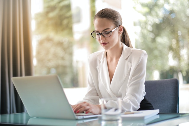 a landlord with black glasses and a blond ponytail types out a rental listing on a white laptop, searching through a department.